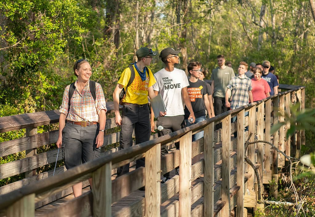 Dr. Watson leading a class of students over a wooden bridge surrounded by trees