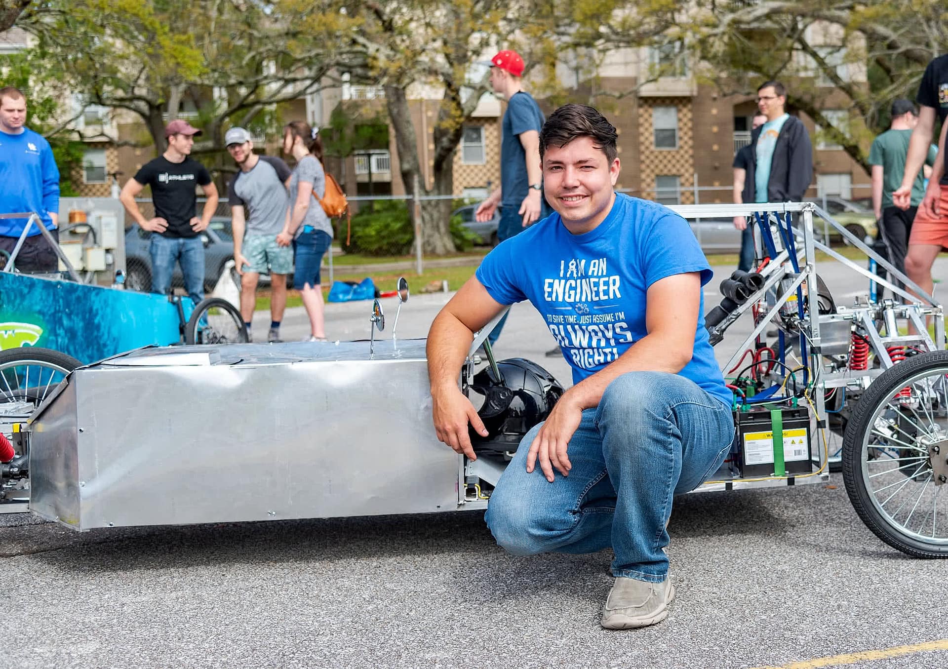 Male student knelt in front of a silver electric vehicle and smiling. 