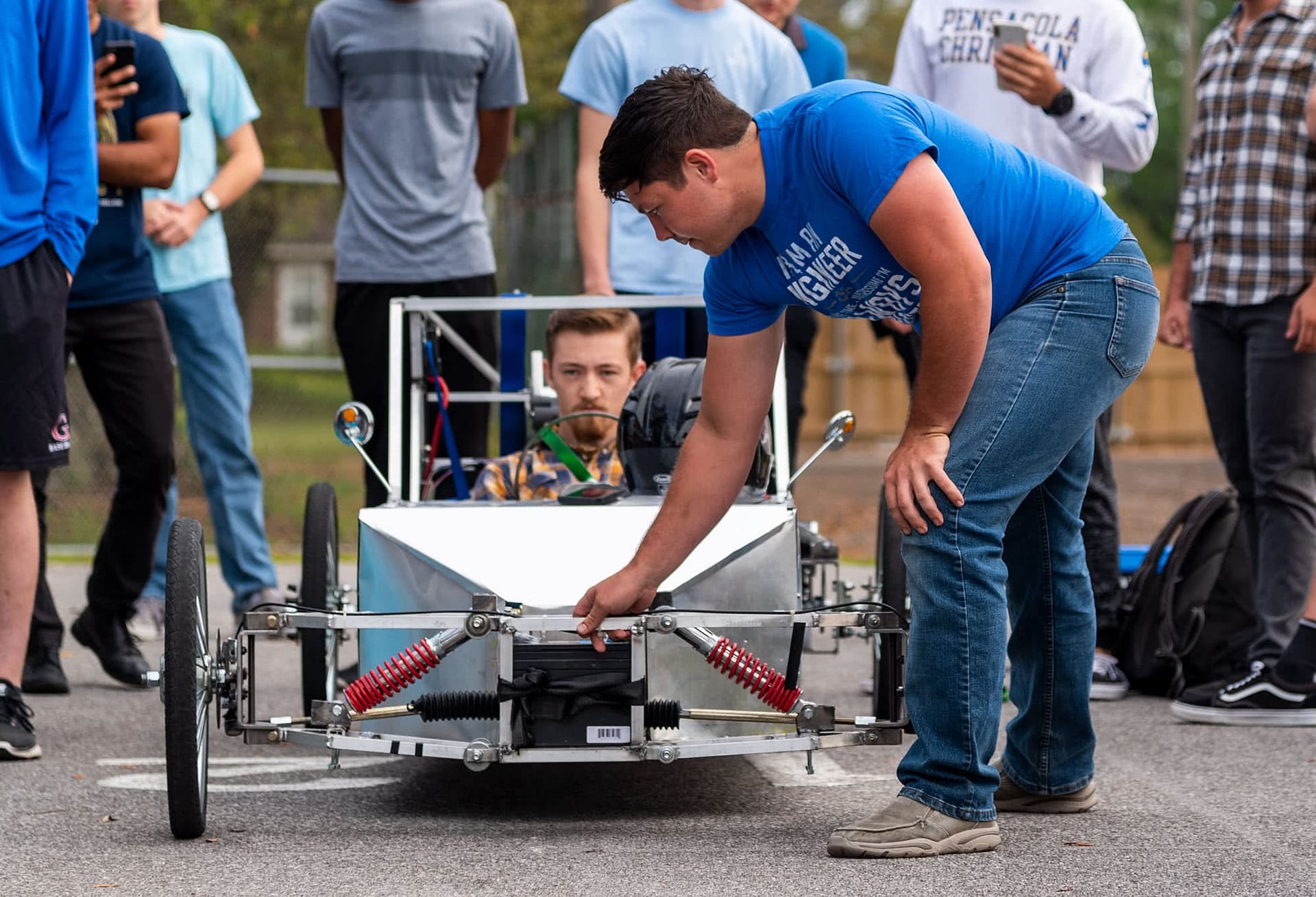 Male student holding the front of an electric vehicle while another male student sits inside the electric vehicle. 