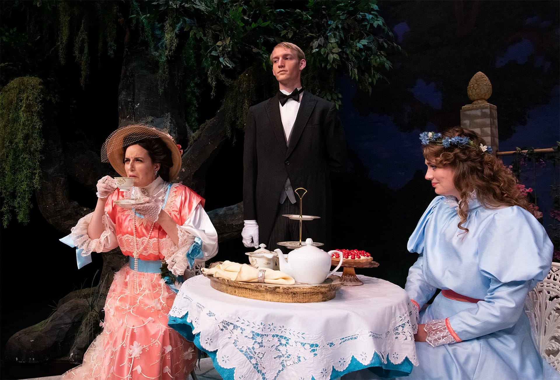 A woman in a blue dress looking at a woman in a pink dress drinking tea. 