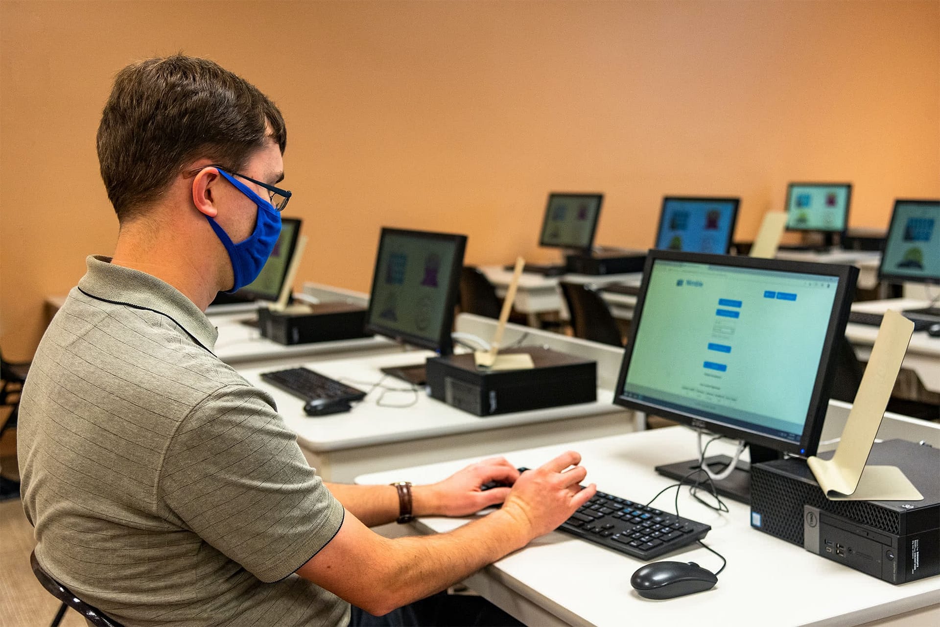 Male student wearing a face covering sits at a computer and types on a keyboard. 
