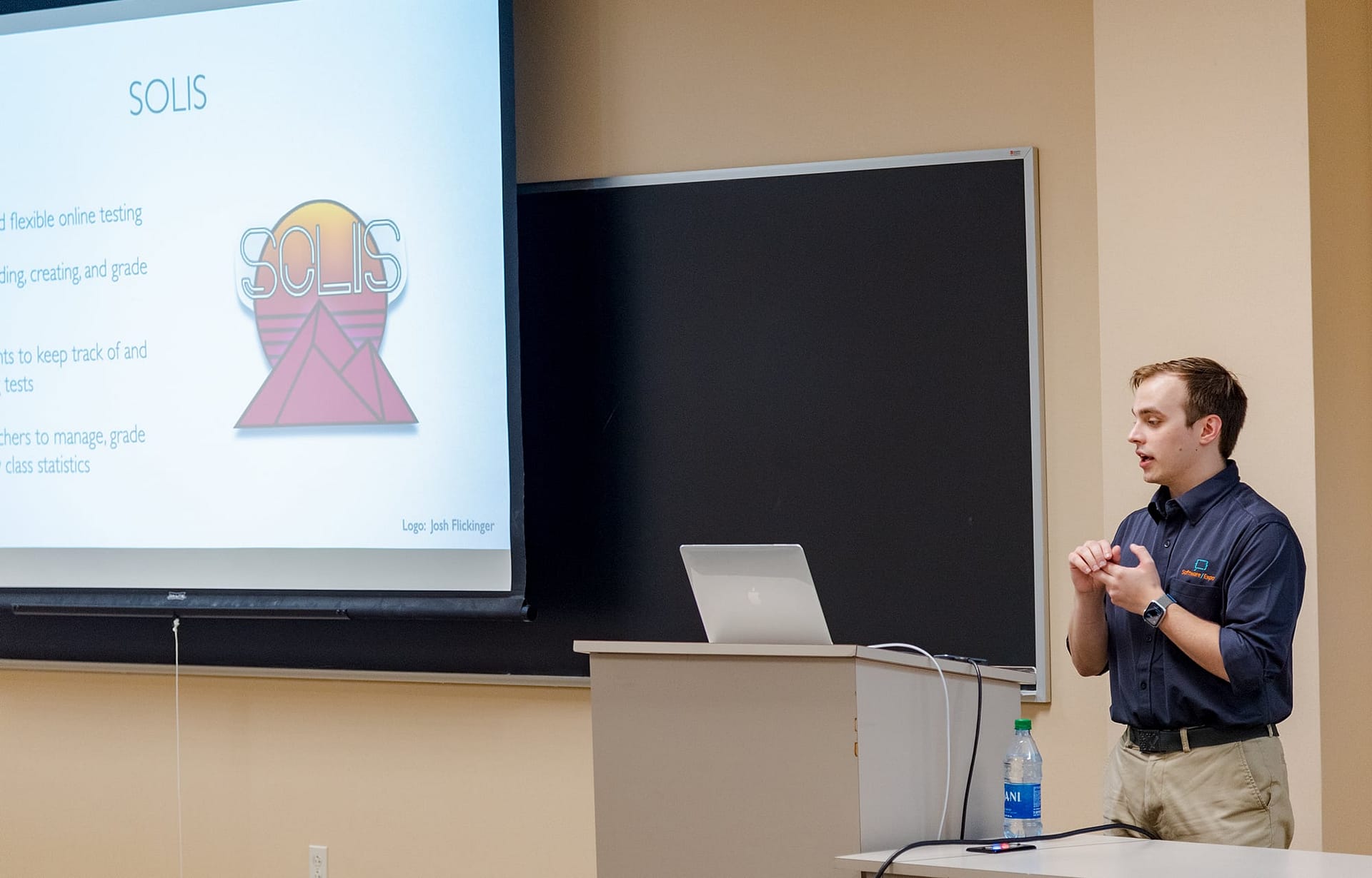 Man standing at the front of a classroom and presenting to a class. 
