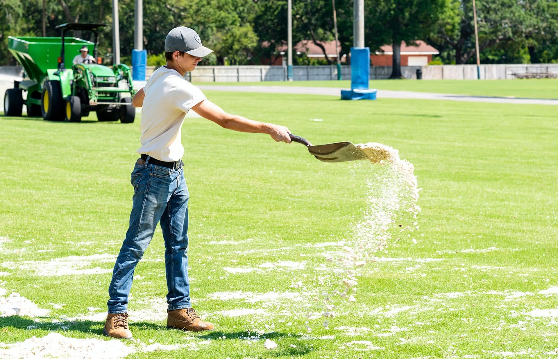 Male student worker spreading sand onto a field with a shovel.
