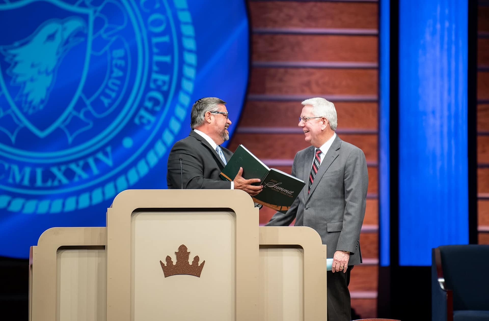 Dr. Shoemaker and Dr. Adkins smiling behind the pulpit with a Summit yearbook in the Crowne Centre at PCC. 