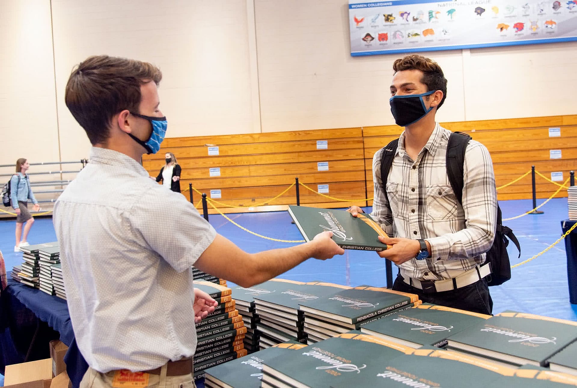Male student handing out a Summit yearbook to another male student wearing a face covering. 