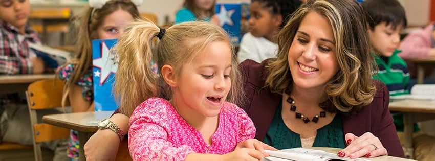 Female teacher smiling and helping female student at the student's desk.