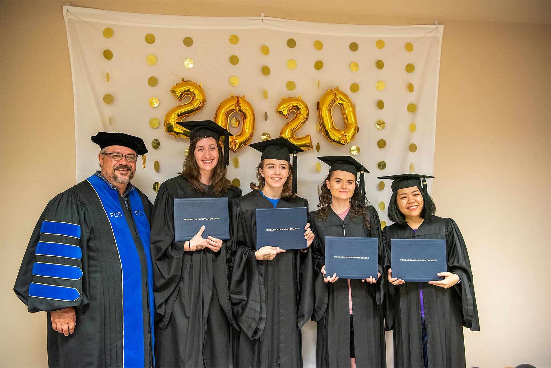 Four PCC graduates in regalia posing with Dr. Shoemaker.