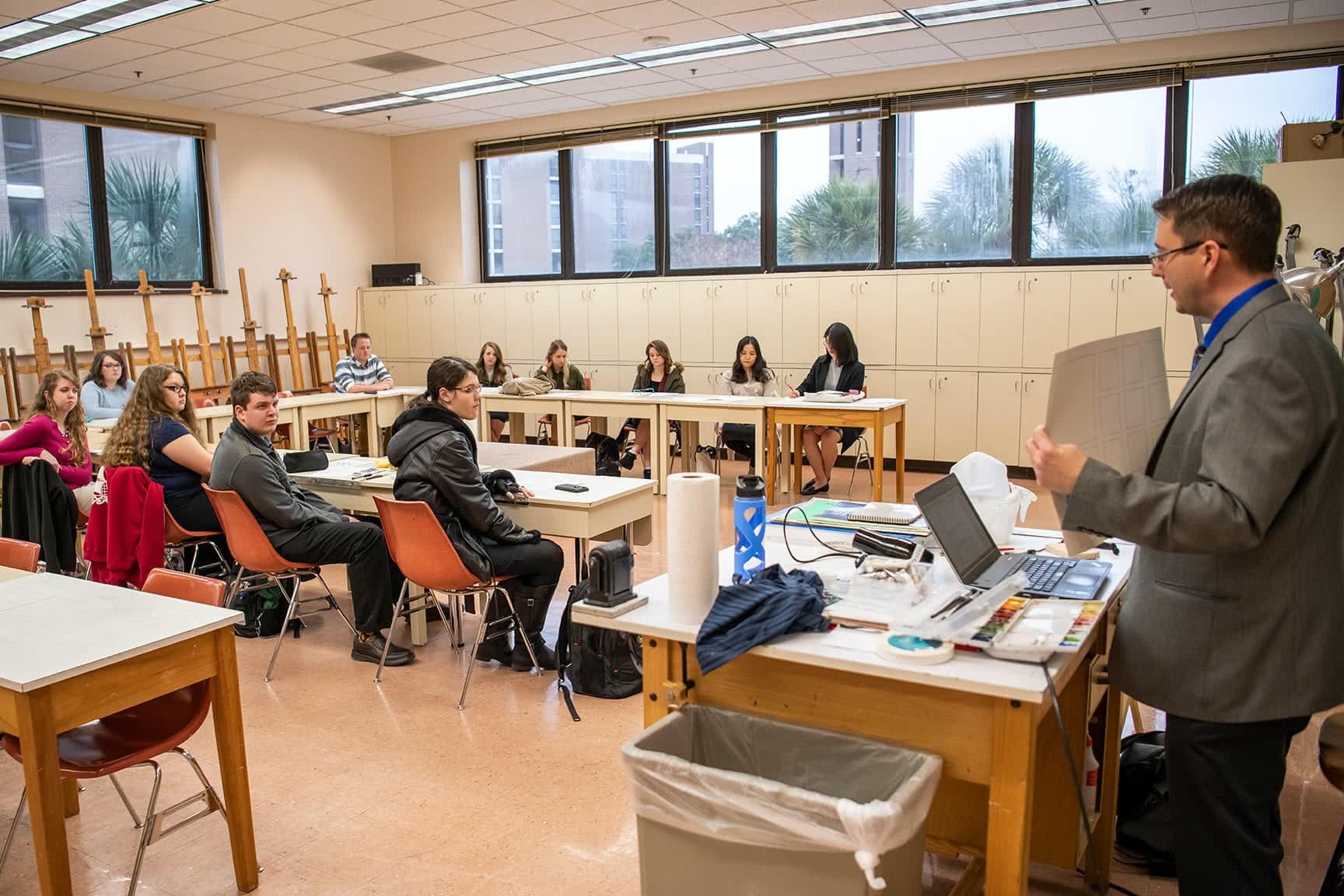 A male teacher holding paper and speaking to a classroom of students sitting at art desks.