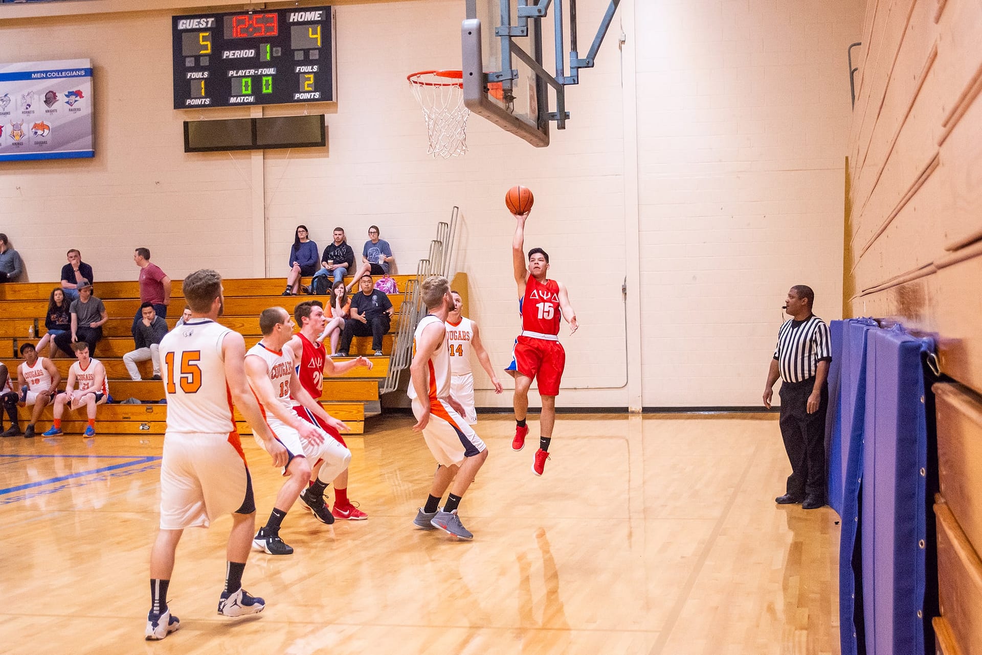 Men's collegian basketball players standing under the net while another player shoots a basketball. 