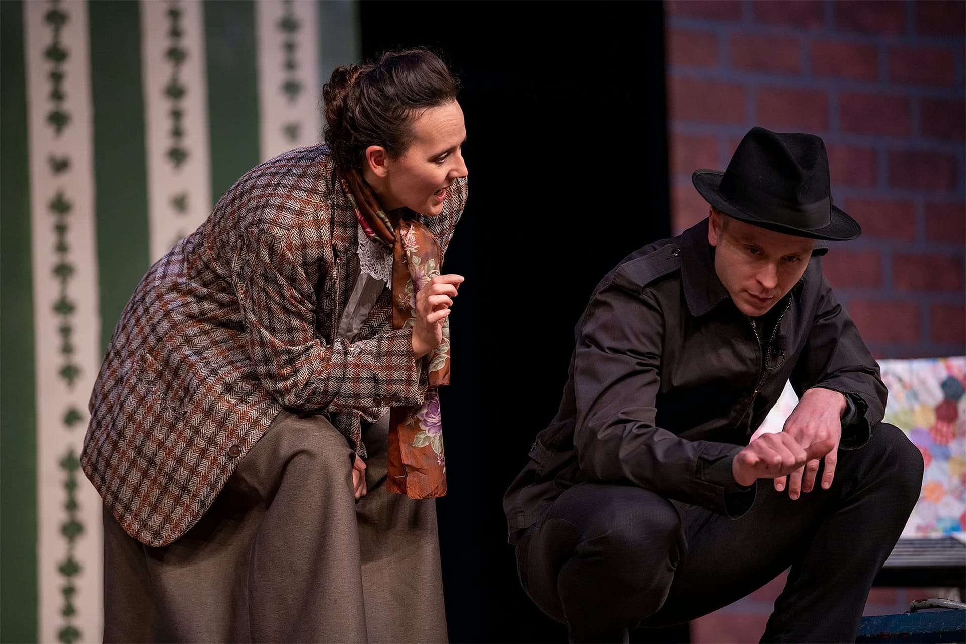 Woman in a plaid jacket and scarf bending over to speak to a man wearing black kneeling down. 