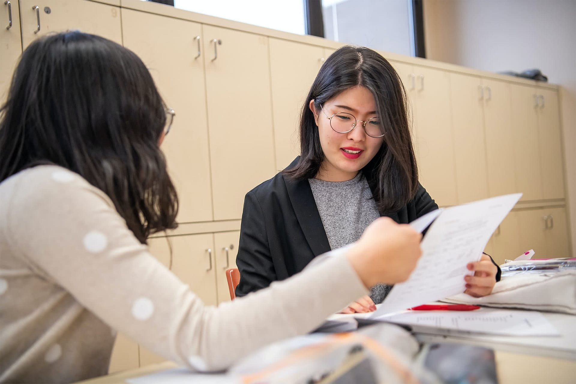 Two female students sitting at art desks and holding papers.
