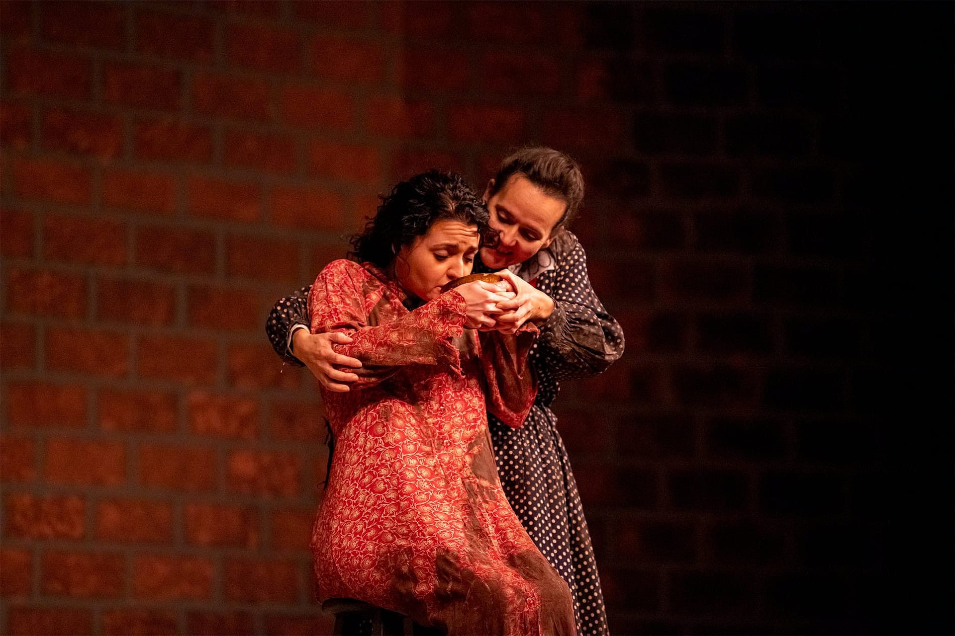 A woman in a black polka dot dress helping a woman in a red dress drink from a bowl. 