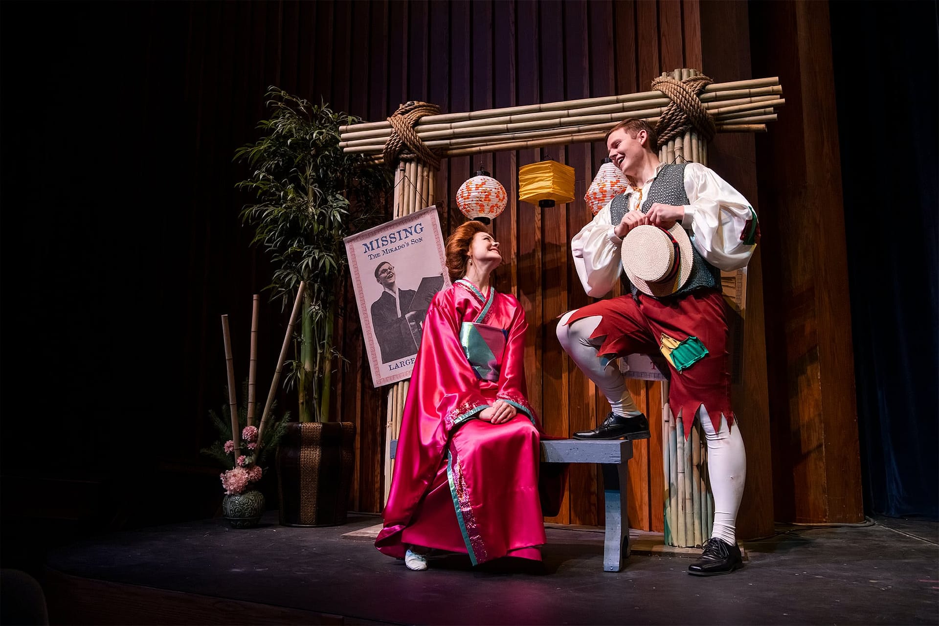 Man wearing a vest and patched pants looking at a woman wearing a pink kimono and sitting on a bench.