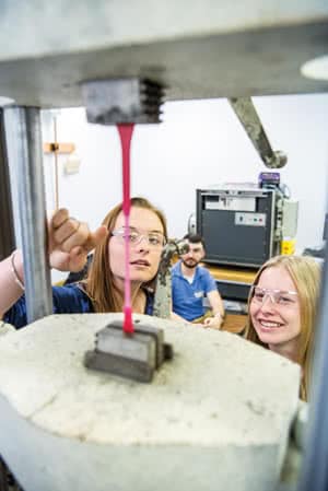 Two female engineering students wearing safety glasses looking at a machine. 