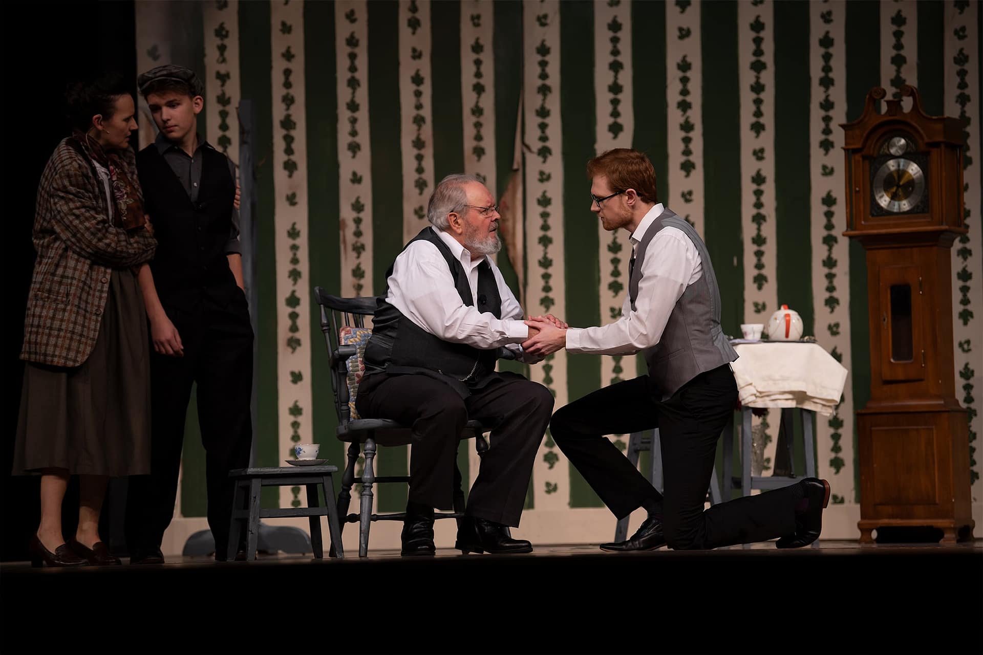 Man wearing a gray vest and glasses on one knee talking to an older man sitting in a chair. 
