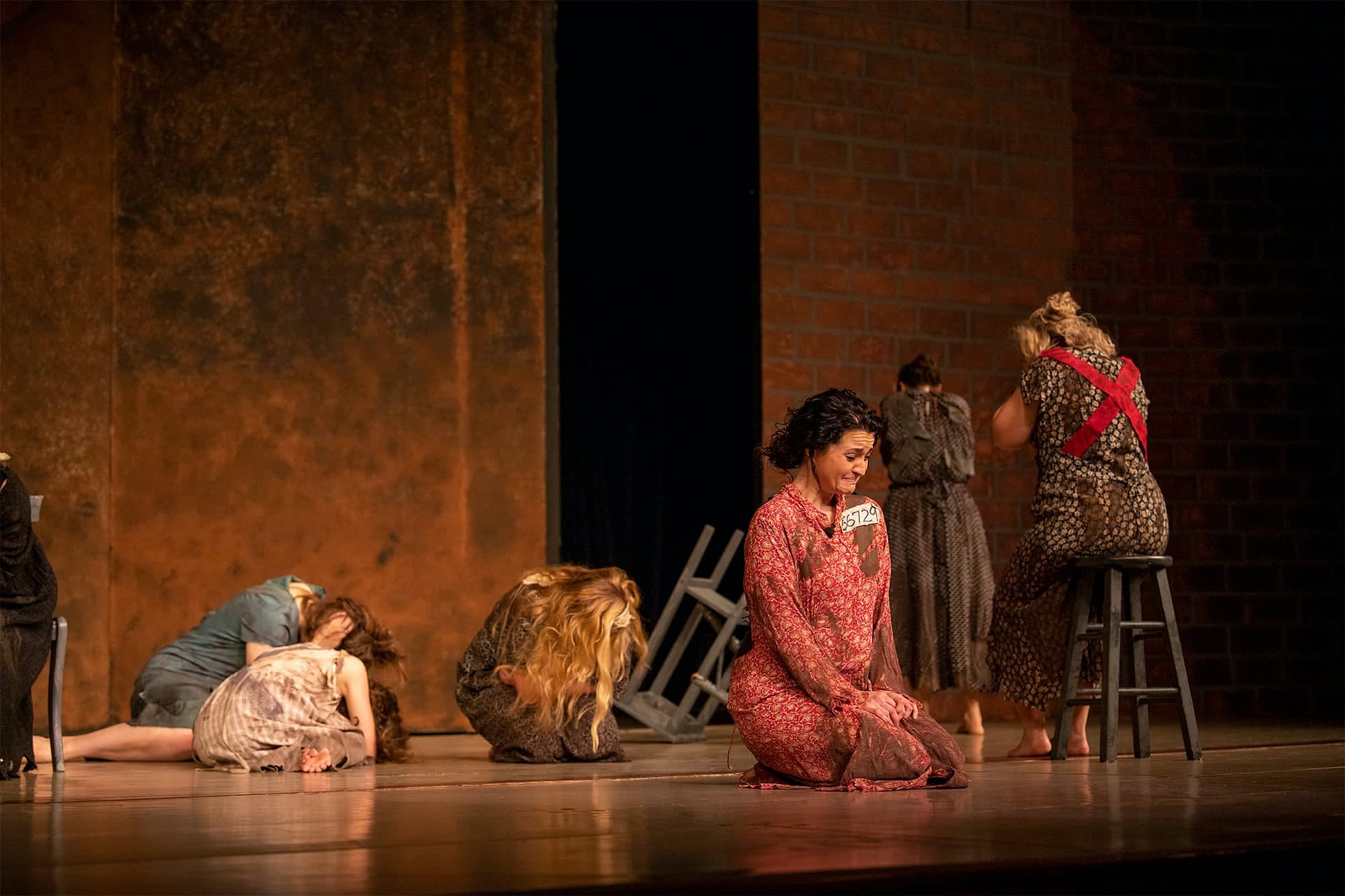 Woman in a pink ragged dress kneeling down while five other women in ragged dresses sit in the background. 