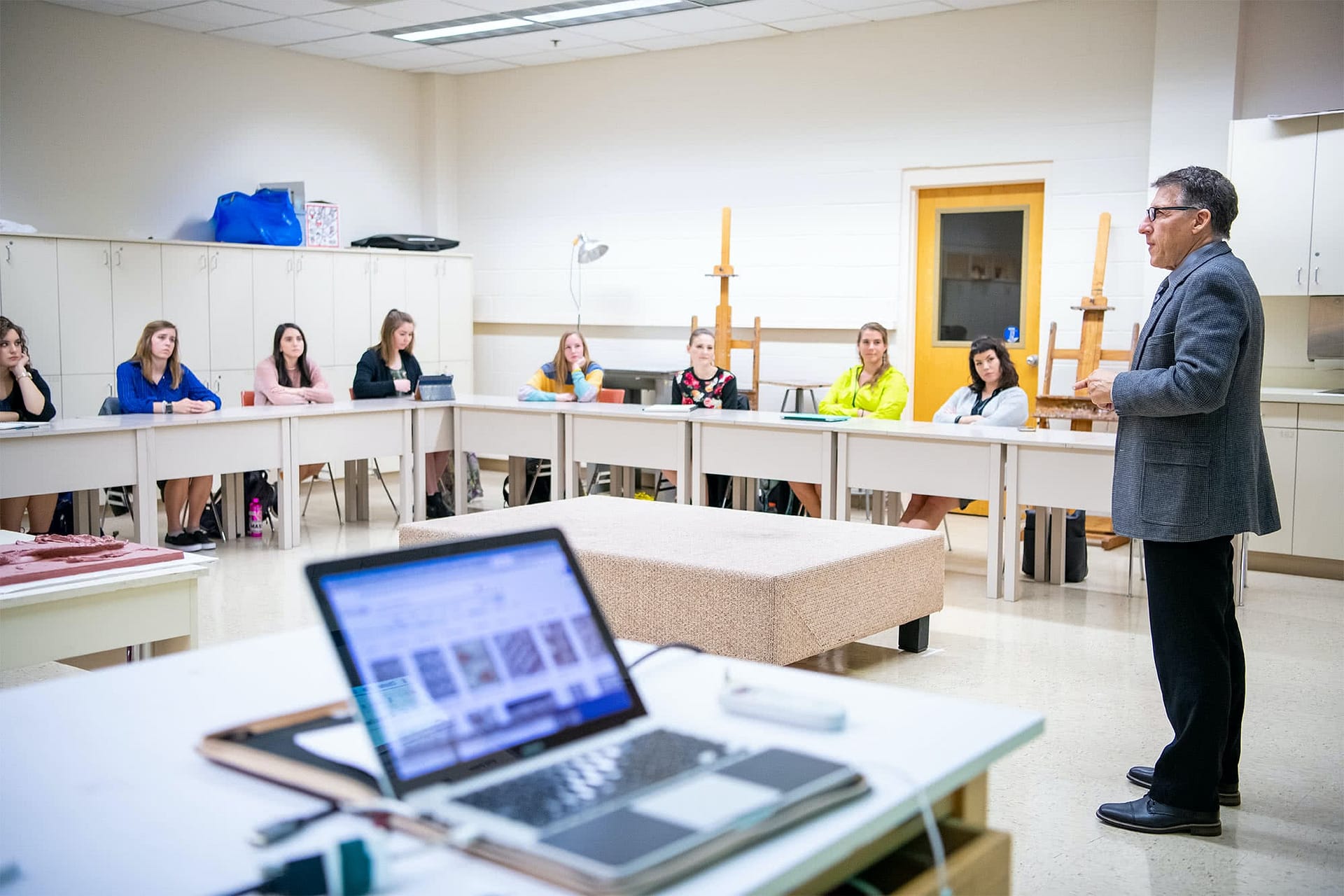 Female students sitting at art desks and listening as their male teacher instructs them.