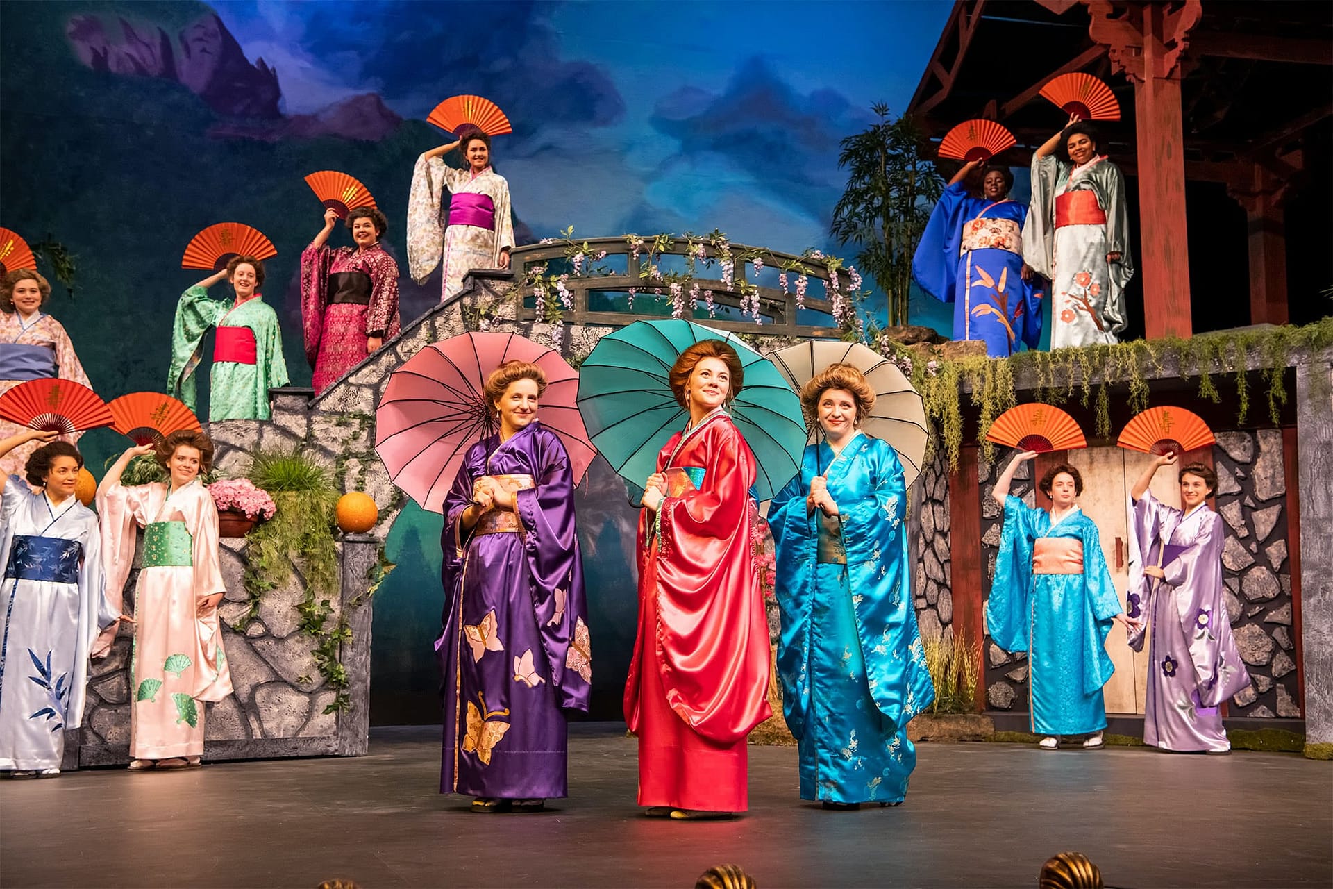 Women holding traditional fans standing and looking at three women wearing colorful kimonos and holding parasols.