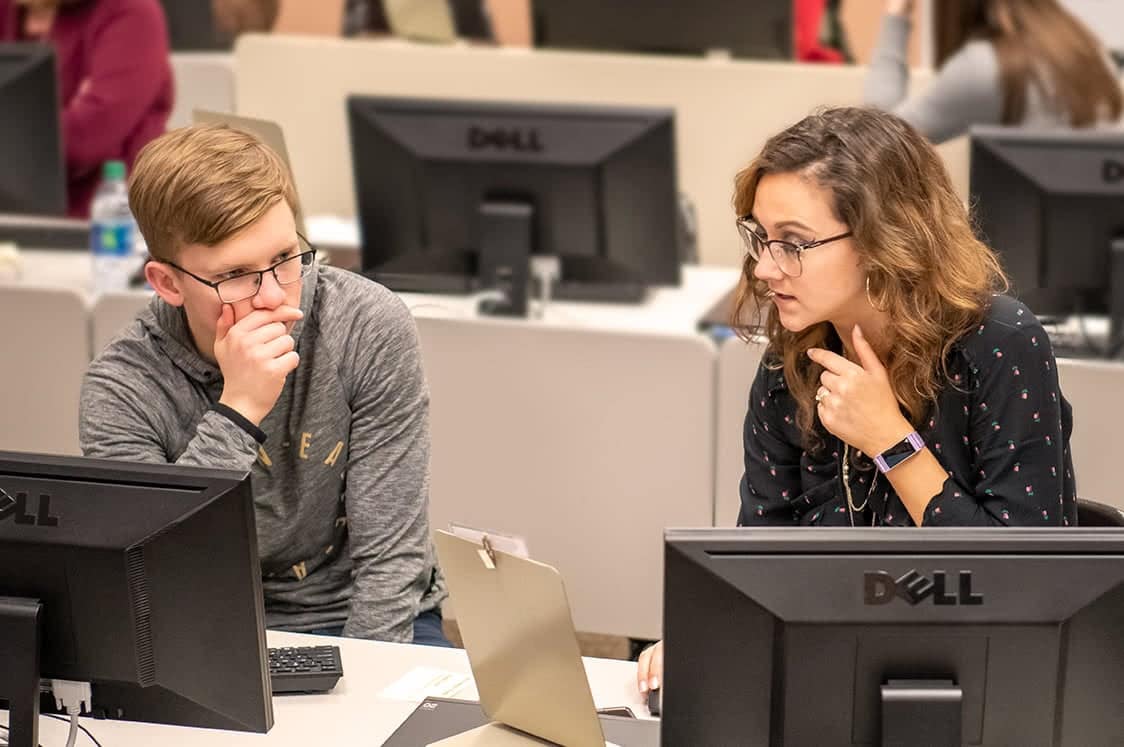 A woman sitting at a computer and helping a male student register for classes for new semester..