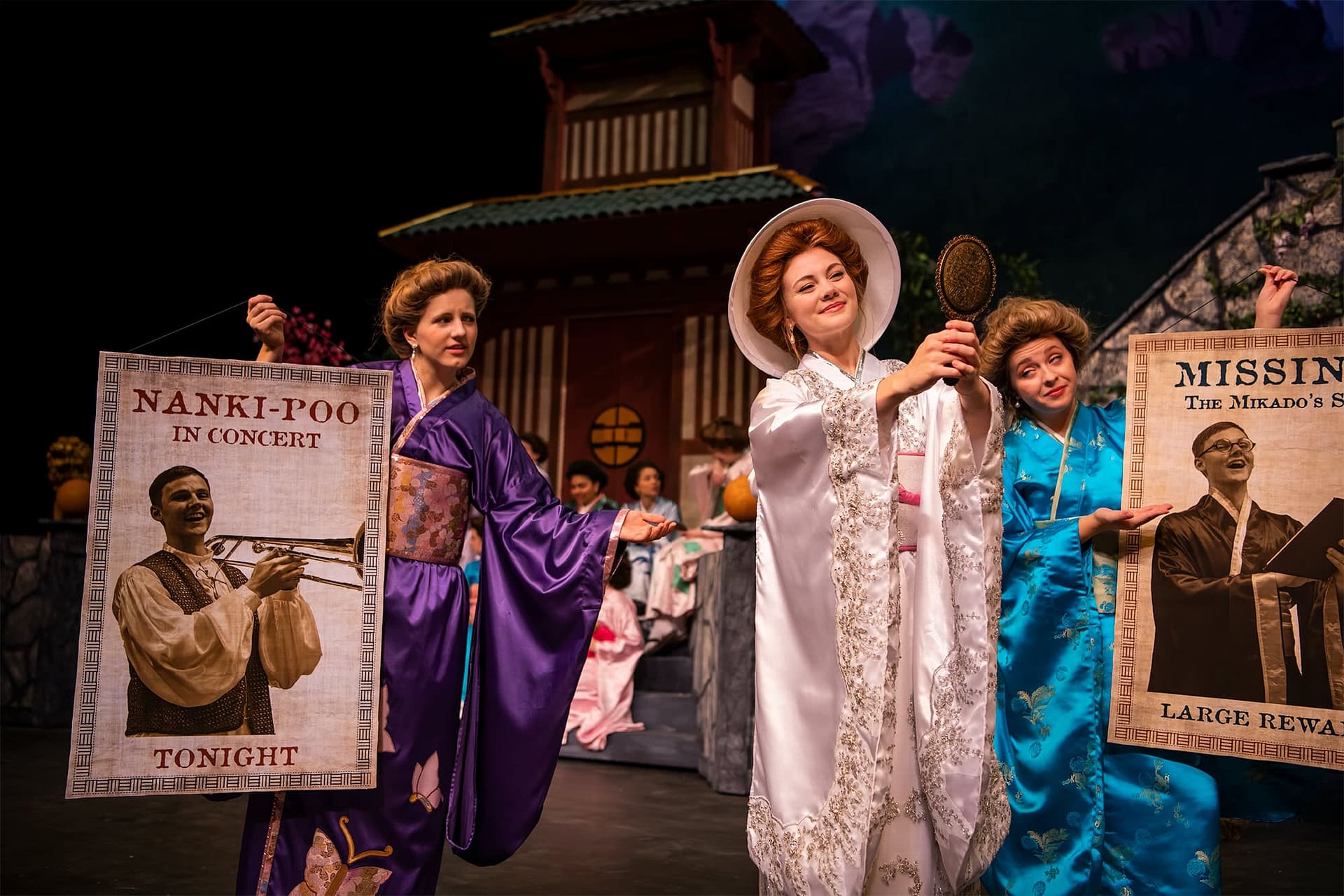 Women in purple and blue kimonos holding signs and looking at a woman wearing white and looking into a mirror.