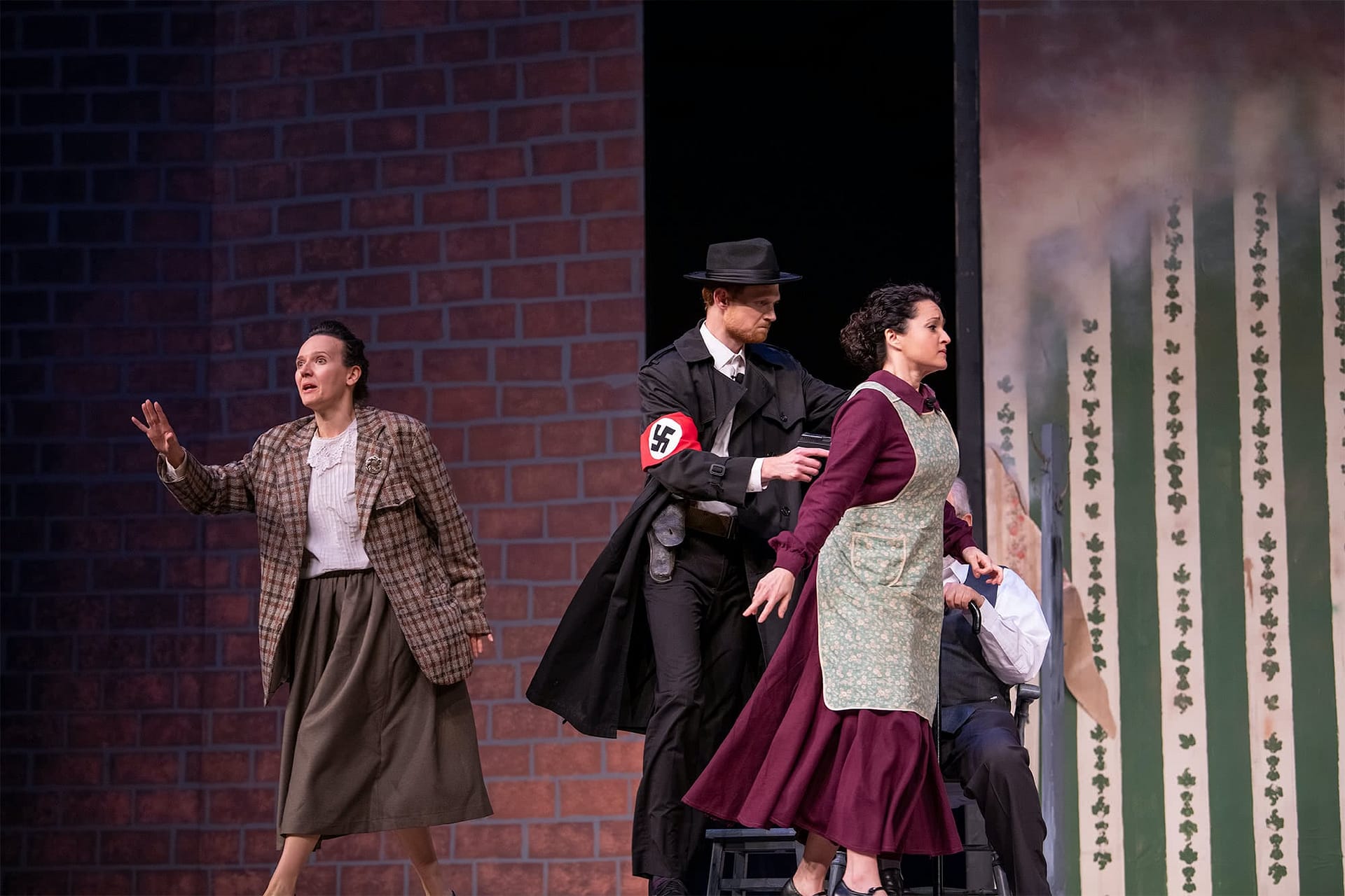 Man in a black suit and hat holding a gun leads a woman wearing an apron away. 