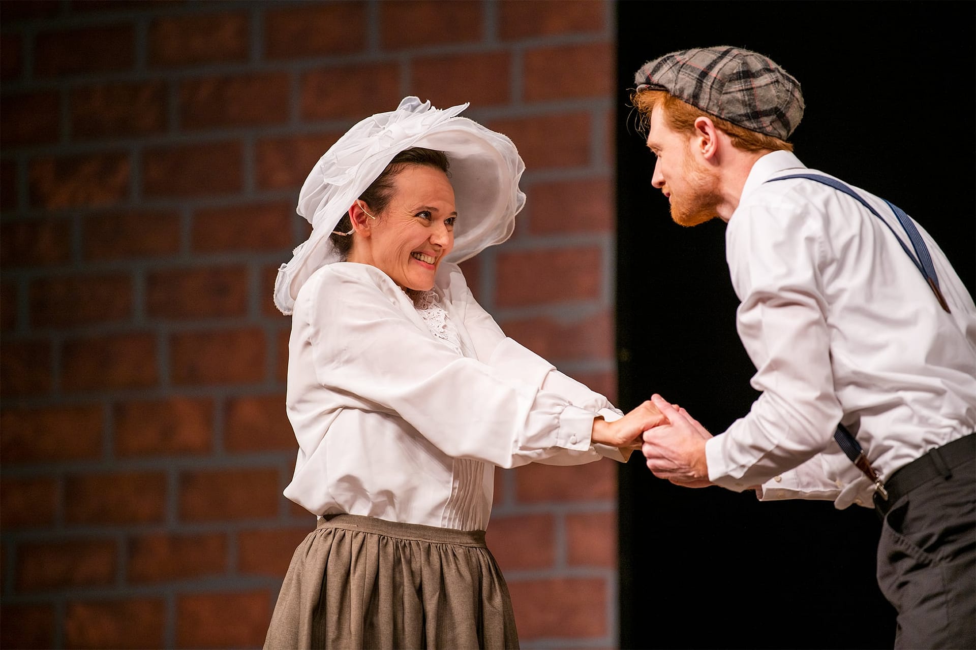A woman in a white hat smiling and holding hands with a man wearing suspenders and a plaid hat. 