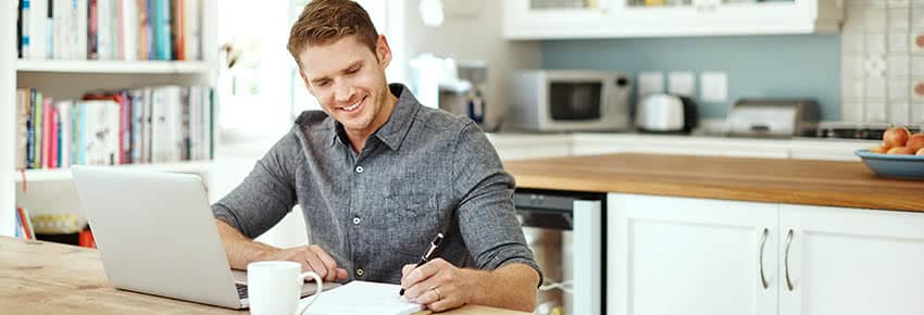 Man sitting at a table with a laptop and writing on a paper.