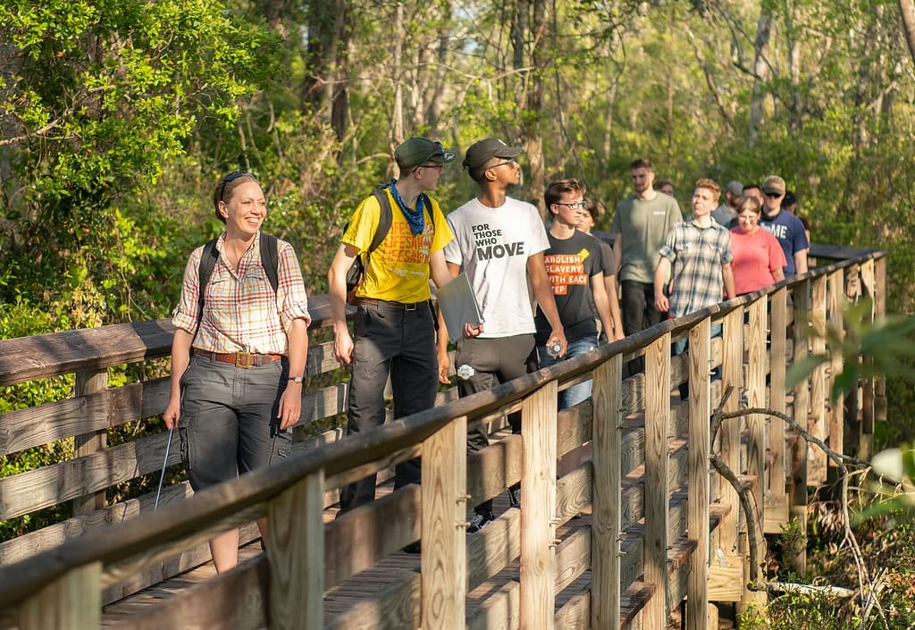 Dr. Watson leading a  class of students over a wooden bridge surrounded by trees