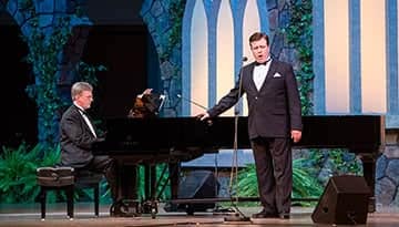 Anthony Kearns sings by the piano, the stage is lit up in blues and greens.