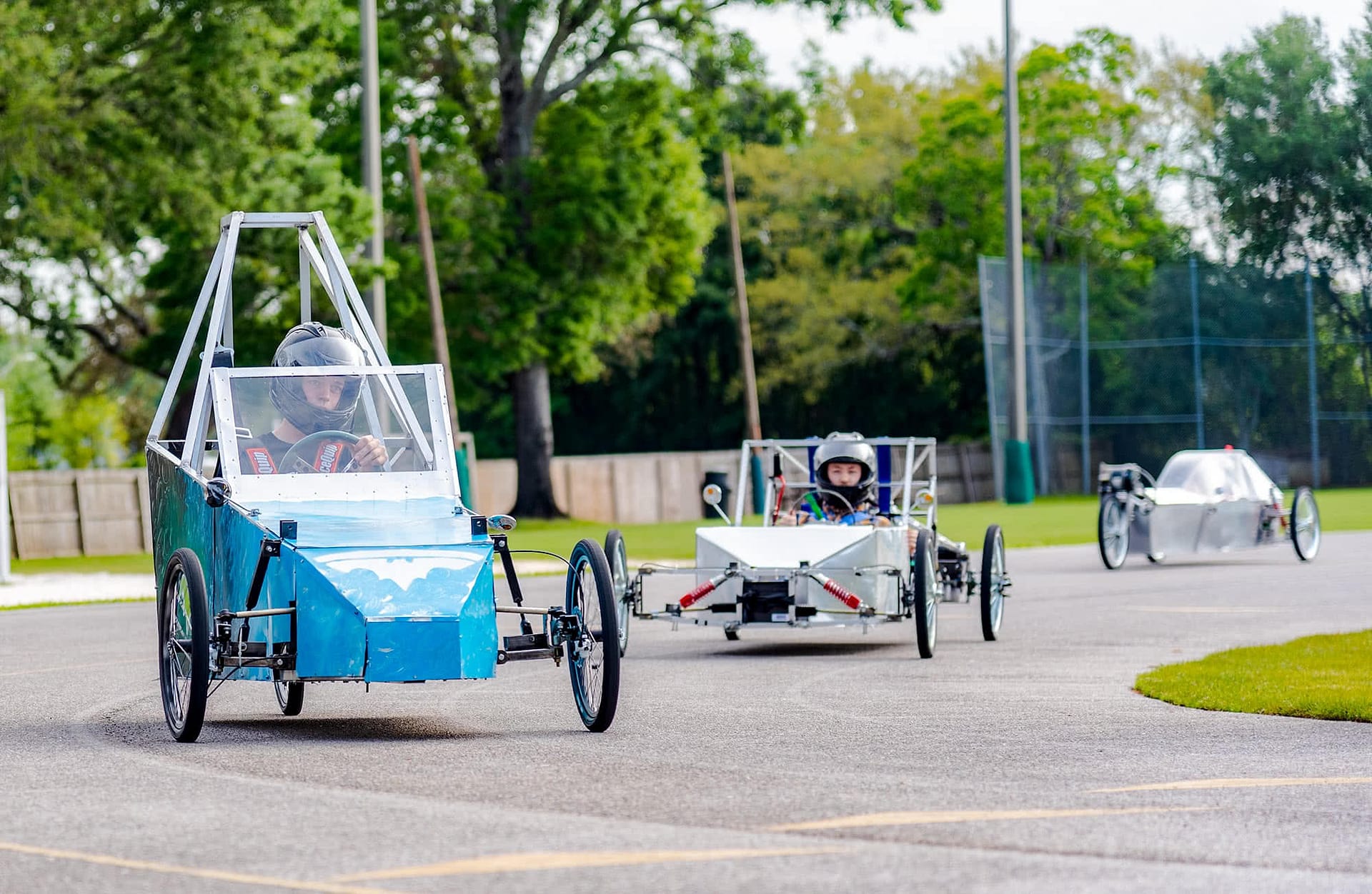 Student in a blue electric vehicle followed by two students in silver electric vehicles. 