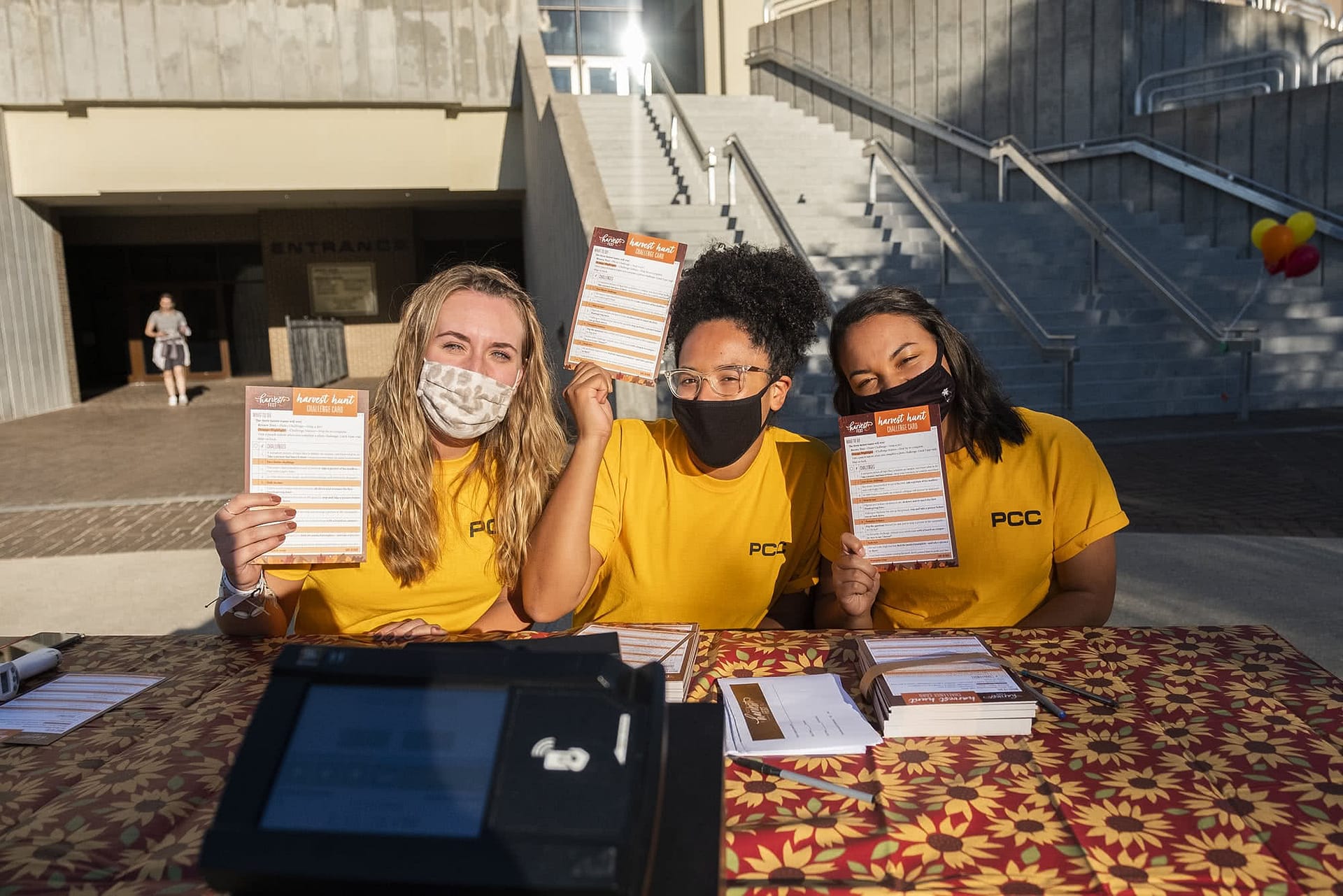 Three female students holding harvest hunt challenge cards at the PCC Harvest Fest. 