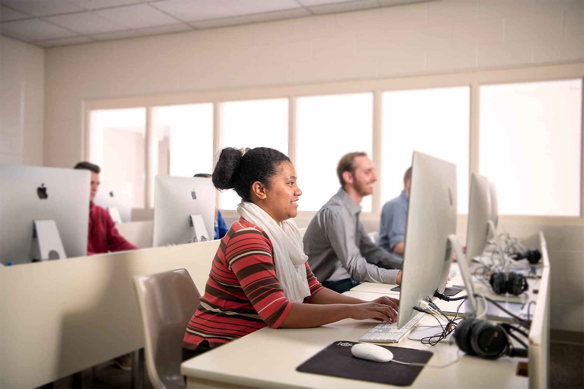 Female interterm student wearing a scarf sitting at a computer with other students in a computer lab.