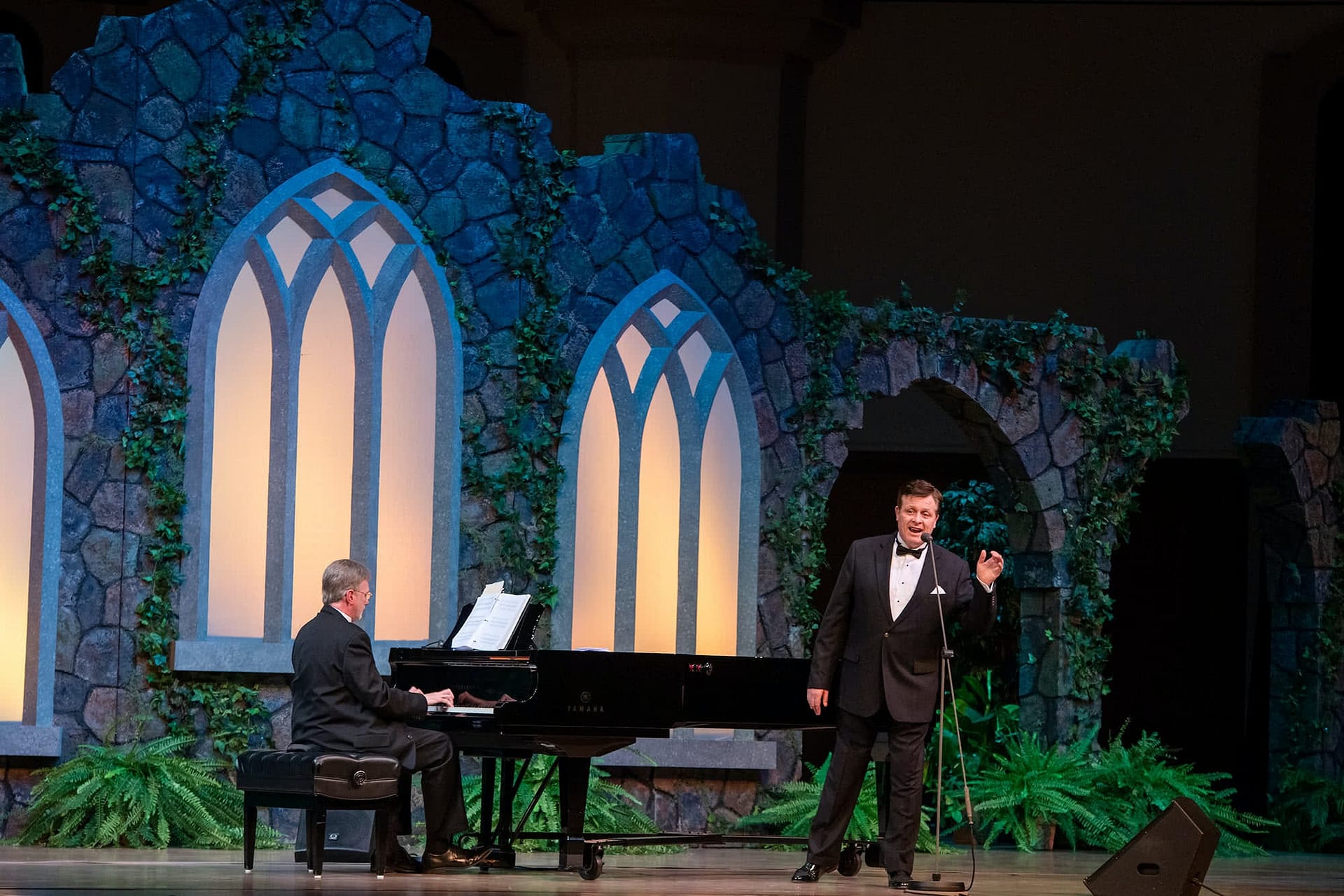 Anthony Kearns sings by the piano, the stage is lit up in blues and greens.