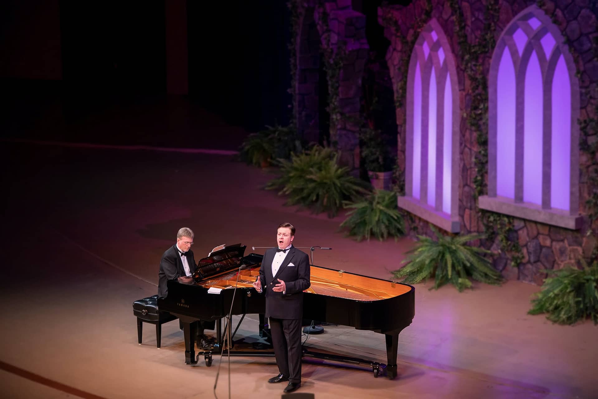 Anthony Kearns sings by the piano, the stage is lit up in purples.