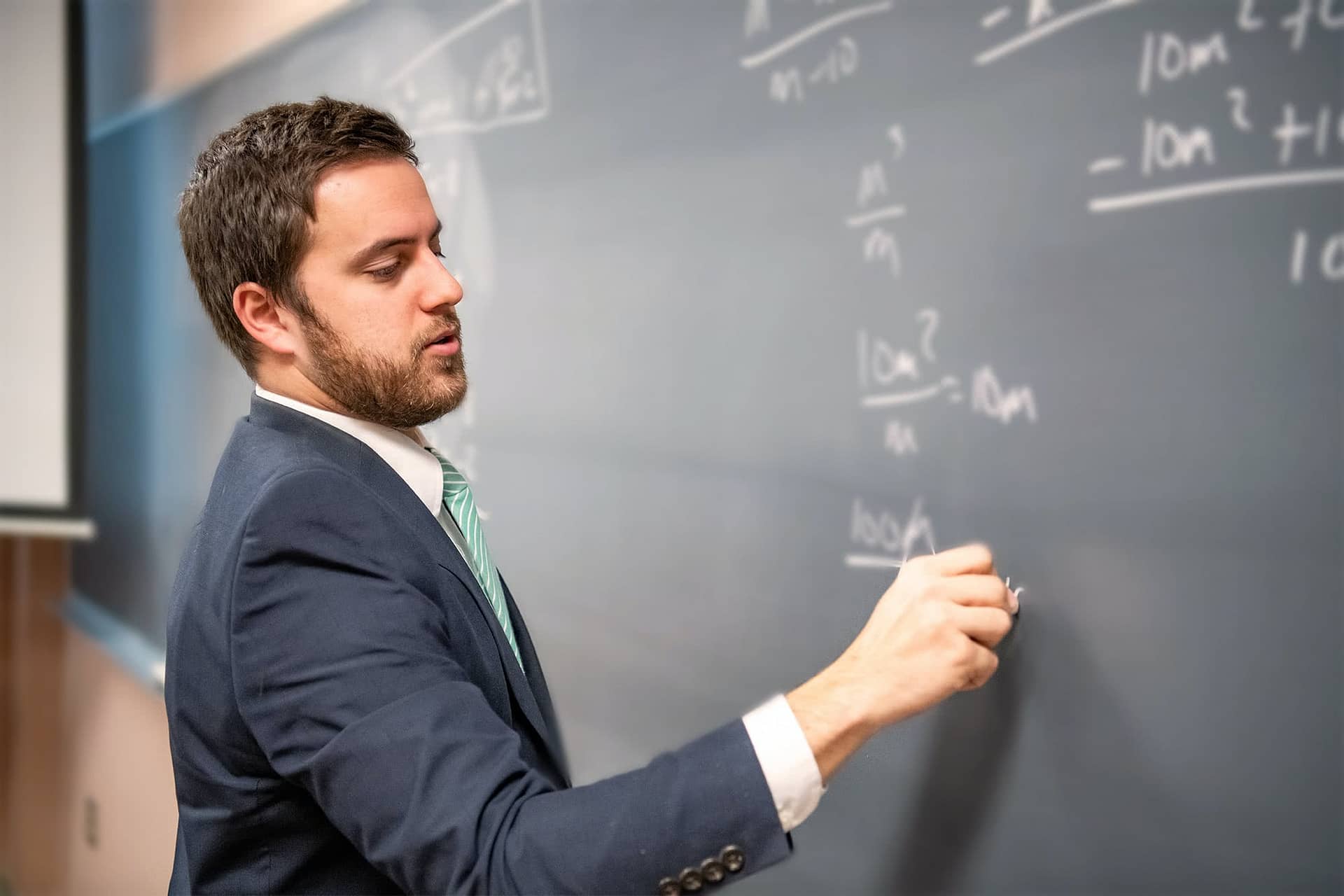 Male interterm teacher writing with chalk on a chalkboard.