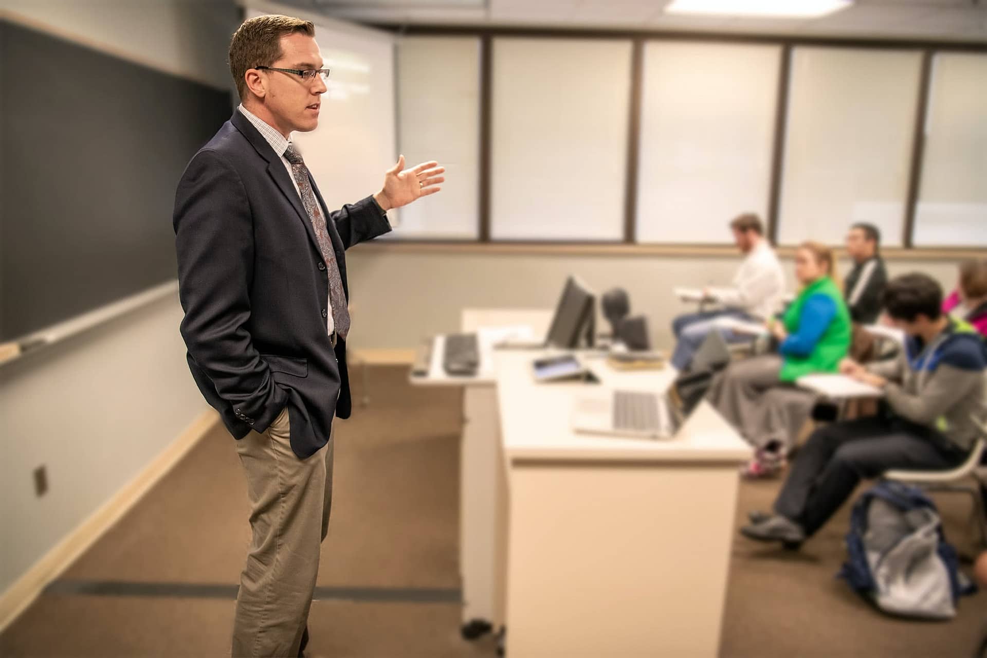 Male interterm teacher standing at the front of a class and speaking to his students.