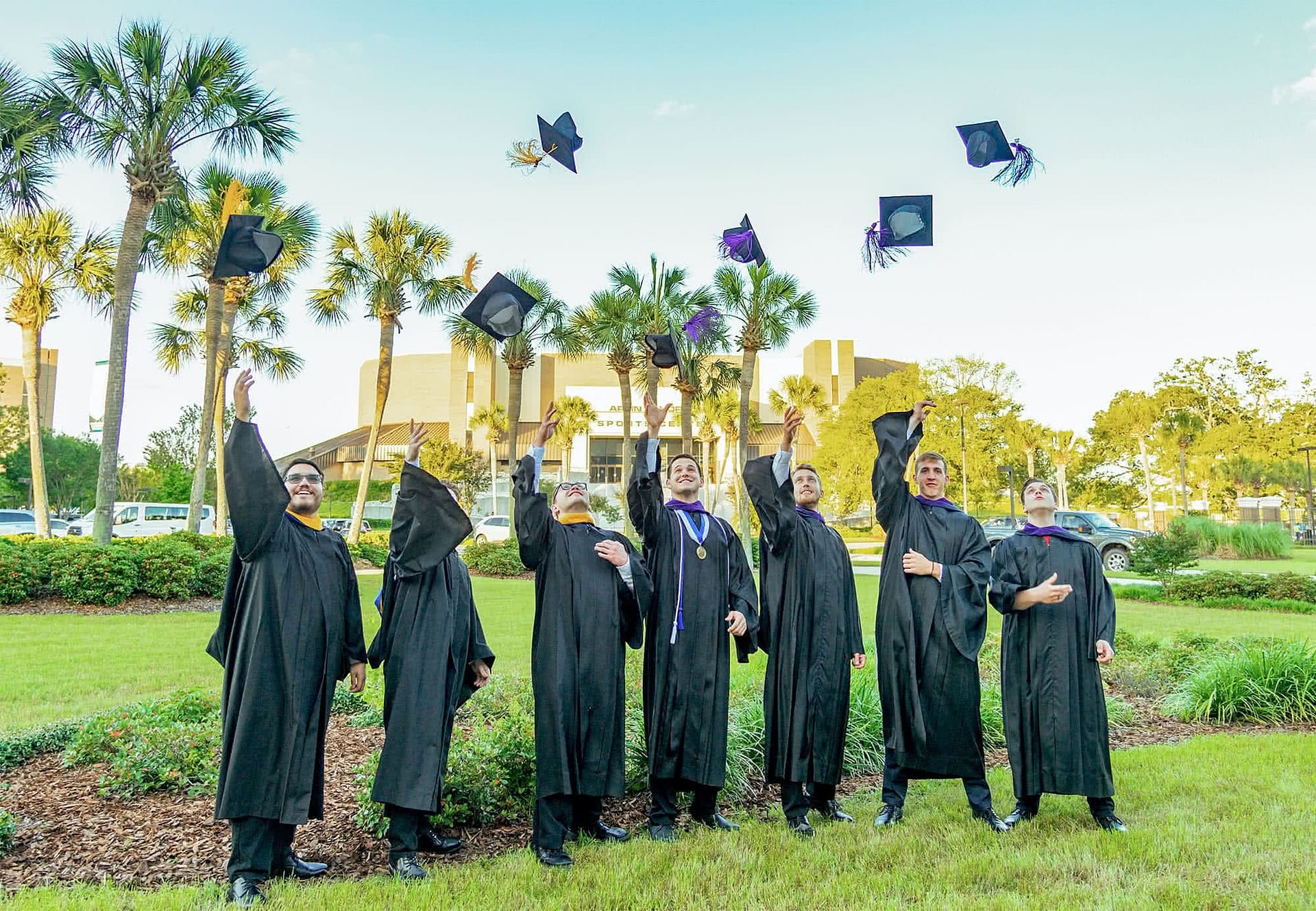 Seven male regalia clad graduates toss caps into the air in front of Sports Center building.